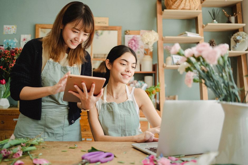 A florist and a customer inside a flower shop, both wearing light green aprons, with the florist holding a tablet and the customer seated at a wooden table. The table displays various pink and white f