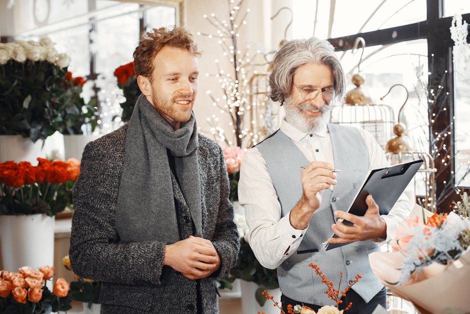 A young man and an older male florist are smiling inside a flower shop filled with vibrant blooms including red, white, and pink flowers arranged in bouquets and vases. The young man, dressed in a dar