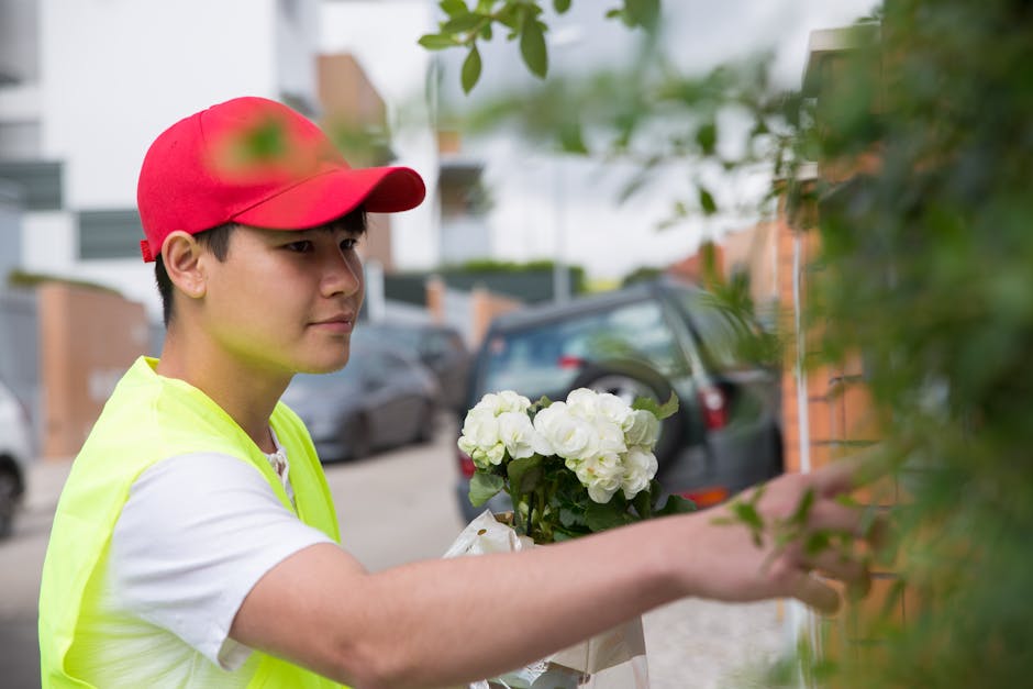 A young delivery person wearing a red cap and a neon yellow safety vest is placing a bouquet of fresh white flowers, including roses and hydrangeas, onto a doorsteps or garden wall in a residential ar