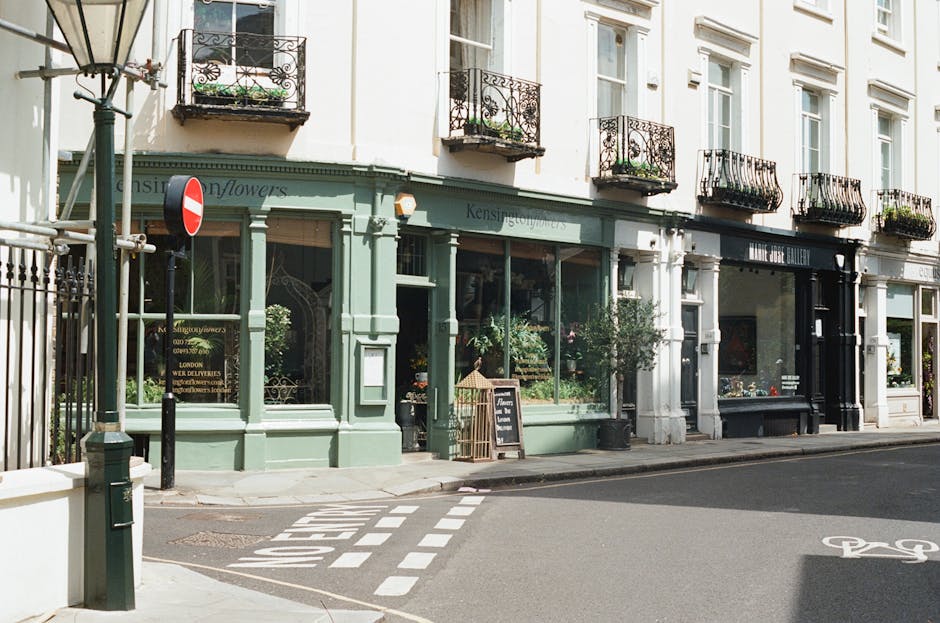 A storefront of a flower shop located on a street corner, painted in light green with large glass windows displaying various floral arrangements inside. The shop has a black signboard with yellow text