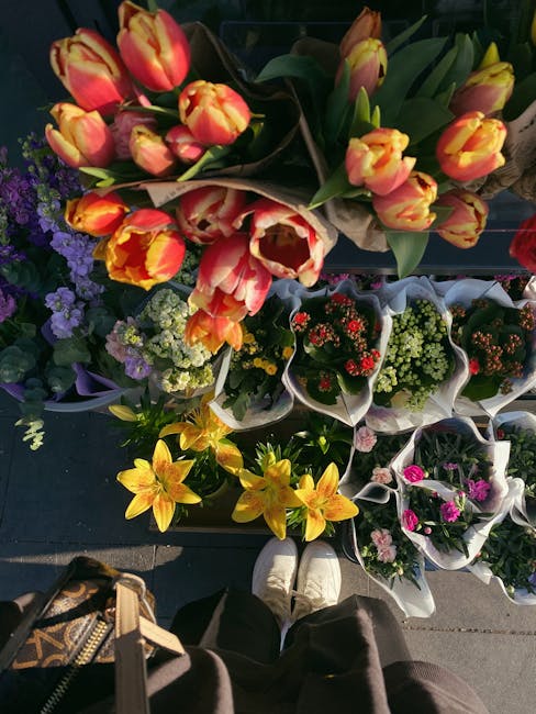 A variety of fresh flower bouquets displayed on a dark surface outdoors, including a prominent arrangement of orange and yellow tulips with green stems and broad leaves, surrounded by smaller potted f