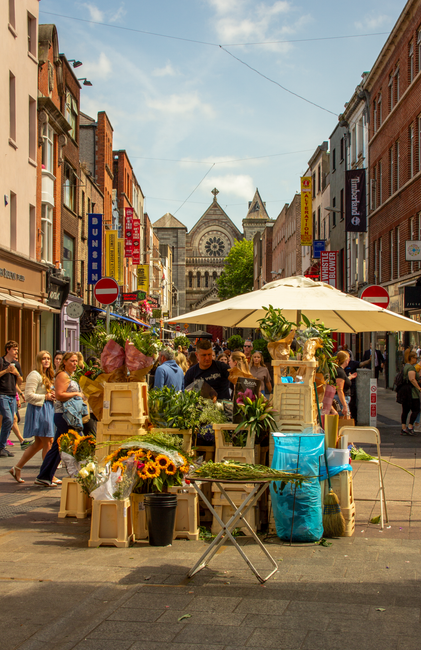 A bustling outdoor street market on Camberwell Church Street with various flower vendors offering fresh floral arrangements. In the foreground, a flower stall displays a variety of blooms, including s
