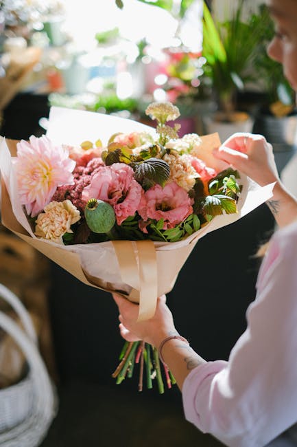 A person holding a large bouquet of fresh flowers, including pink peonies, white carnations, and purple statice, wrapped in light brown craft paper and tied with a cream satin ribbon. The bouquet feat