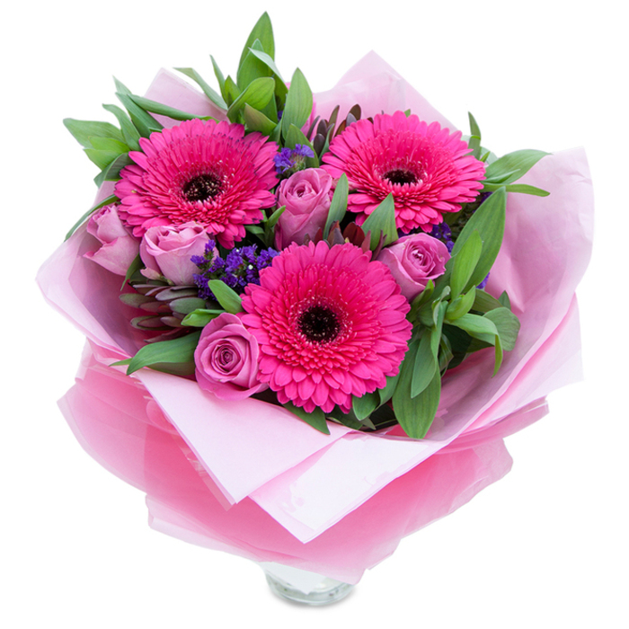 There is an almost garden-like charm held within this carefully arranged bunch: three fuchsia Gerbera daisies stand wide-eyed and proud, their layered petals forming strong, graphic circles around dark, almost inked centres that give the bouquet visual weight. Surrounding them, roses in a muted lavender-pink or dusty rose unfurl in softer, more intimate spirals, their silky petals catching a faint, powdery fragrance. Flecks of deep violet from small statice clusters thread through the composition, adding delicate, papery texture and the suggestion of wild hedgerow blossoms tucked into a cultivated spray. The greenery - broad, textured leaves that recall salal or eucalyptus - is placed with intentional spacing, giving breathing room to each bloom while creating a lush, verdant backdrop that reads as both elegant and freshly cut. The flowers are enveloped in a layer of soft pale pink tissue and an outer wrap of translucent fuchsia-pink cellophane, slightly crinkled to reflect light and to emphasise the bouquet's rosy theme; the wrapping is folded with the careful hand of a florist. At the lower edge the rim of a clear glass vase is visible, indicating the arrangement is ready to be displayed at a bedside, in a study, or as a centrepiece. I imagine this piece brightening a table on Camberwell Church Street or bringing color to a living room near Burgess Park - a composed, garden-inflected bouquet that speaks of quiet celebration and considered craftsmanship.