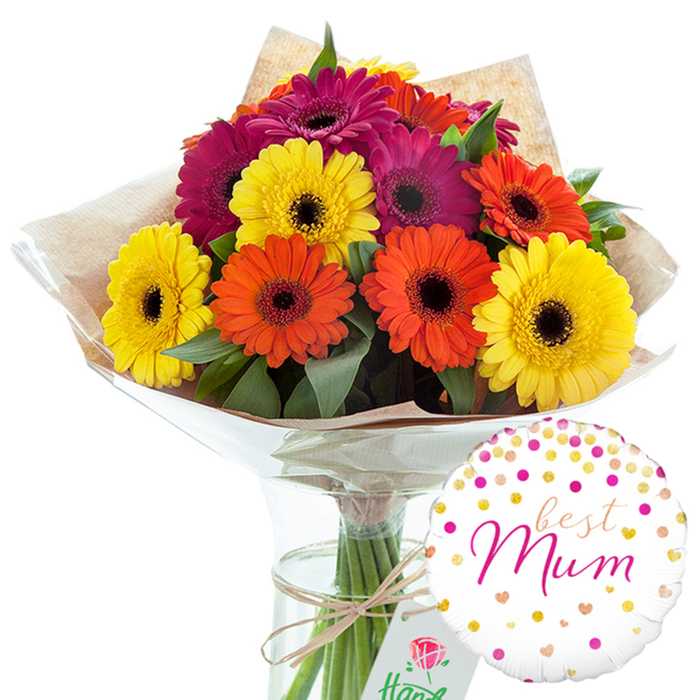 The photograph presents a joyful floral gift set, composed with the calm precision of an experienced Camberwell florist preparing an order for same-day delivery. At the centre is a lush bouquet of Gerbera daisies, the blooms arranged in a loose dome that almost overflows the rim of a tall, clear glass vase. The daisies are large and immaculate, each flower fully open to reveal an inky dark central disk encircled by rings of tightly packed petals. Their colours move in a harmonious trio: bright lemon yellow, vibrant orange reminiscent of a late sunset over Camberwell Green, and a rich magenta that leans toward deep fuchsia. Pockets of glossy, dark green leaves appear between the blooms, softening the bright palette and adding a sense of garden freshness, as though the flowers were gathered from a well-kept back garden off Coldharbour Lane. The stems are bound together at the base and swaddled in gently creased brown kraft paper, which gives a tactile, rustic contrast to the smooth petals. Around this sits a sheath of clear, reflective cellophane that protects the arrangement while allowing every colour and texture to remain visible. A simple tie of natural string or twine secures the wrapping, its small, hand-tied bow suggesting care rather than extravagance. The vase's transparent walls reveal the straight, green stems and the water line, emphasising the bouquet's freshness. To the lower right, a round white helium balloon leans toward the flowers, its surface dotted with scattered confetti-like circles and hearts in gold and various pinks. At its centre, the words best Mum appear in sweeping pink calligraphy, making the image unmistakably about celebrating a mother, whether for Mother's Day, a birthday, or a special thank-you from someone in SE5.